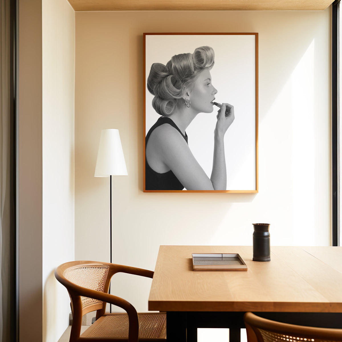 Woman in hair rollers applying lipstick profile print in an oak frame above a dining table and cane chair