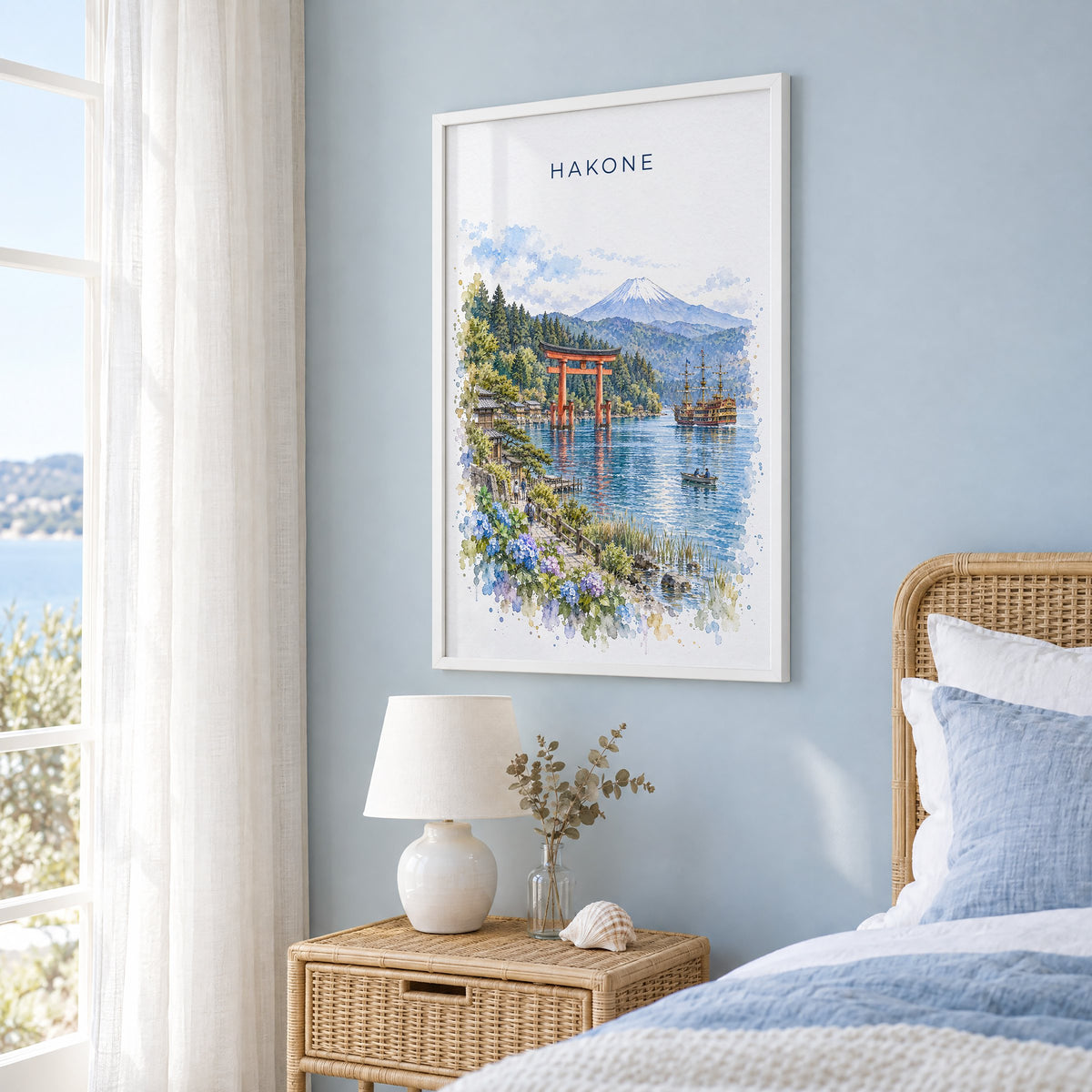 Hakone watercolor print in a white frame on a pale blue wall beside a rattan bed in a coastal bedroom