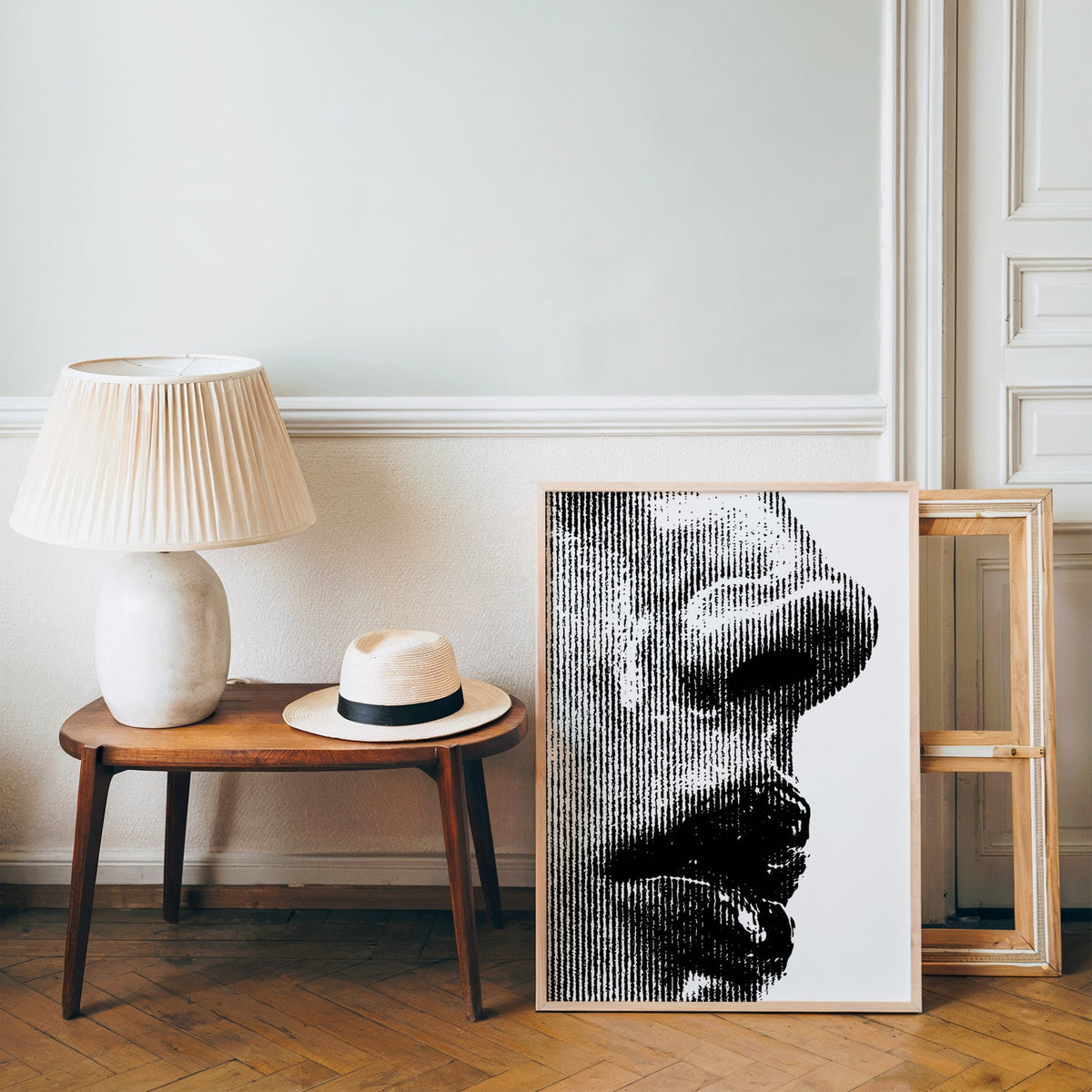 Halftone line face profile graphic print in a pine frame on a wood floor beside a lamp and straw hat