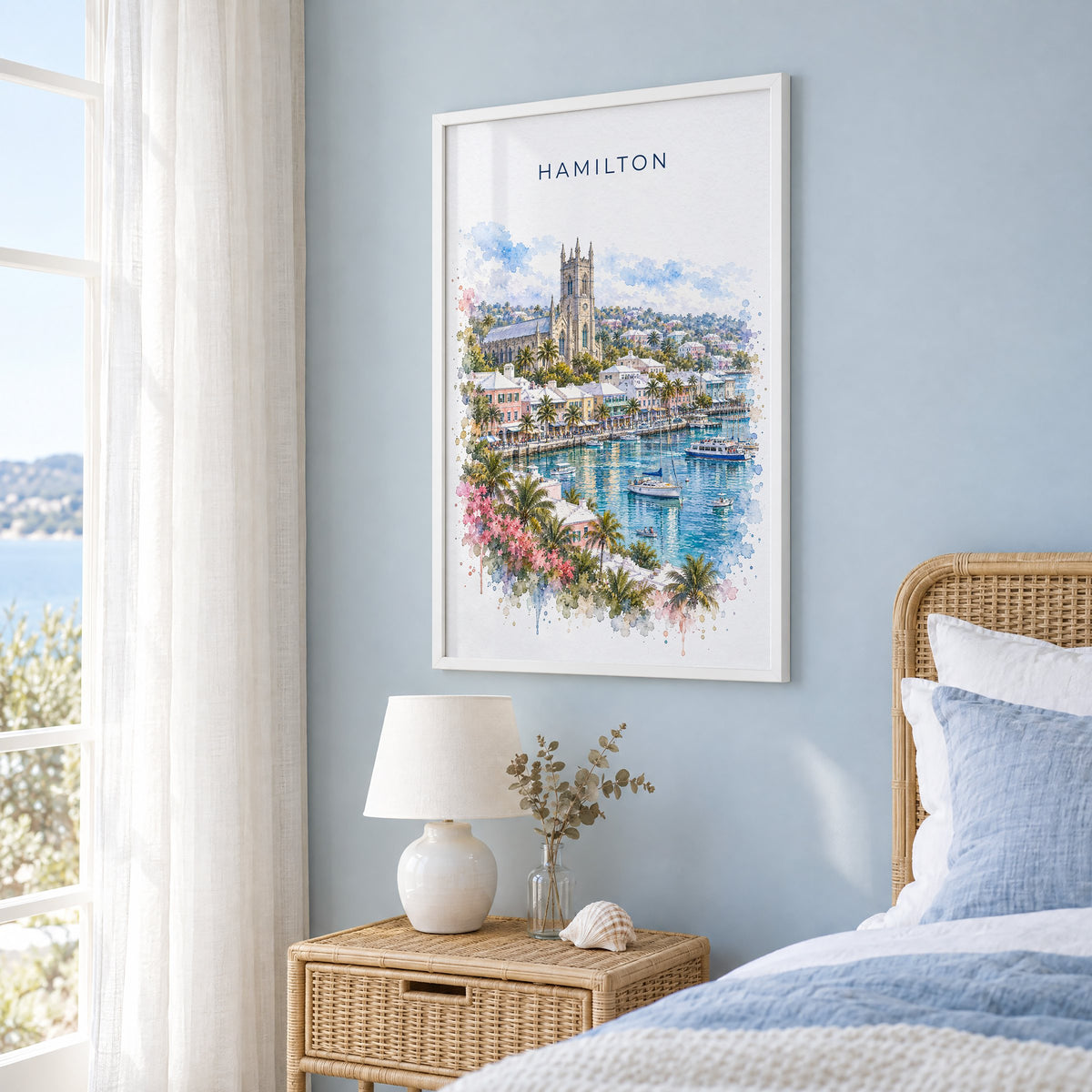 Hamilton, Bermuda watercolor print in a white frame on a pale blue wall beside a rattan bed in a coastal bedroom