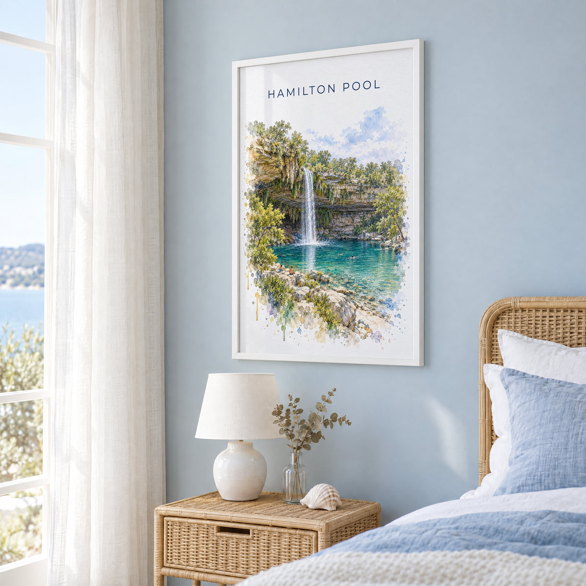 Hamilton Pool watercolor print in a white frame on a pale blue wall beside a rattan bed in a coastal bedroom