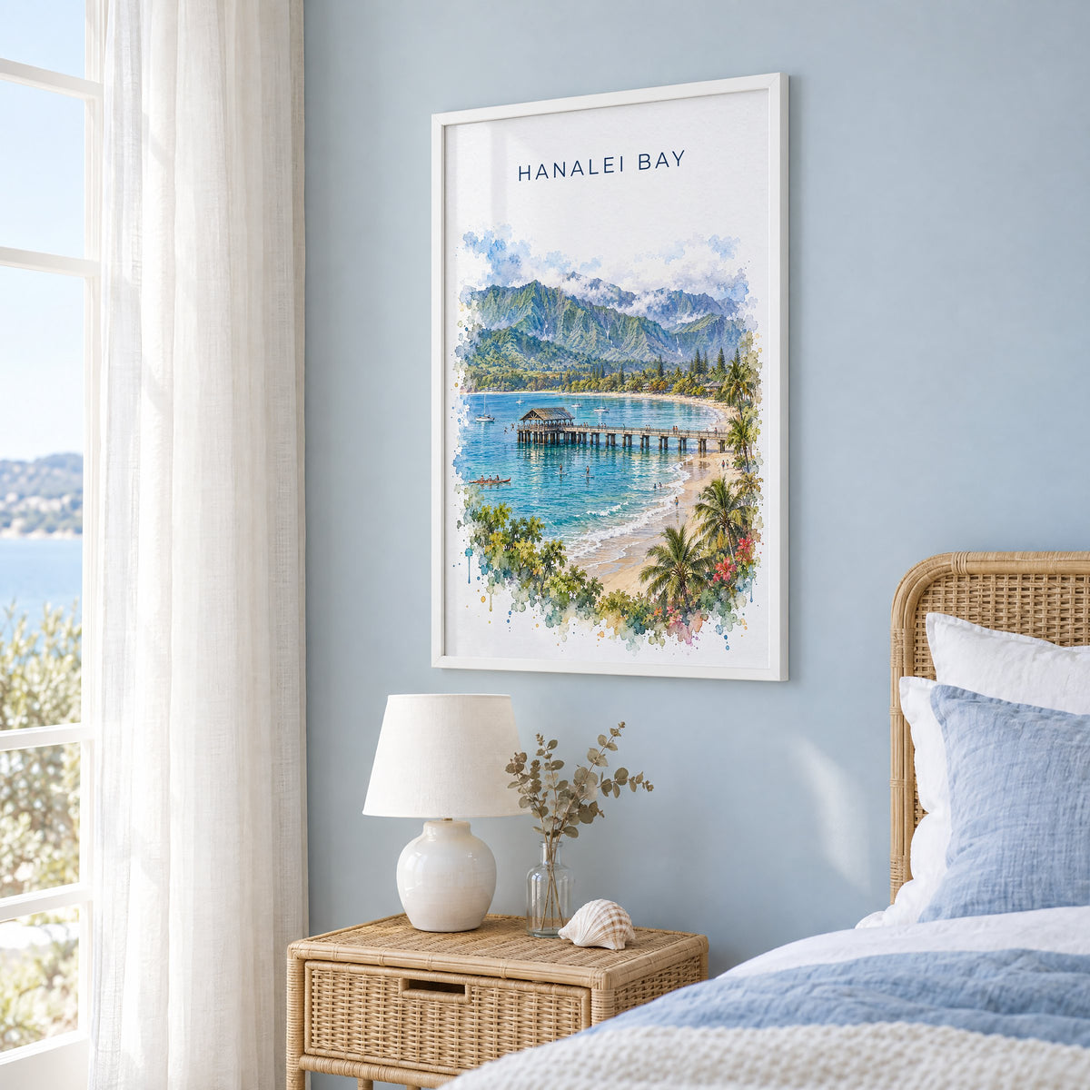 Hanalei Bay watercolor print in a white frame on a pale blue wall beside a rattan bed in a coastal bedroom