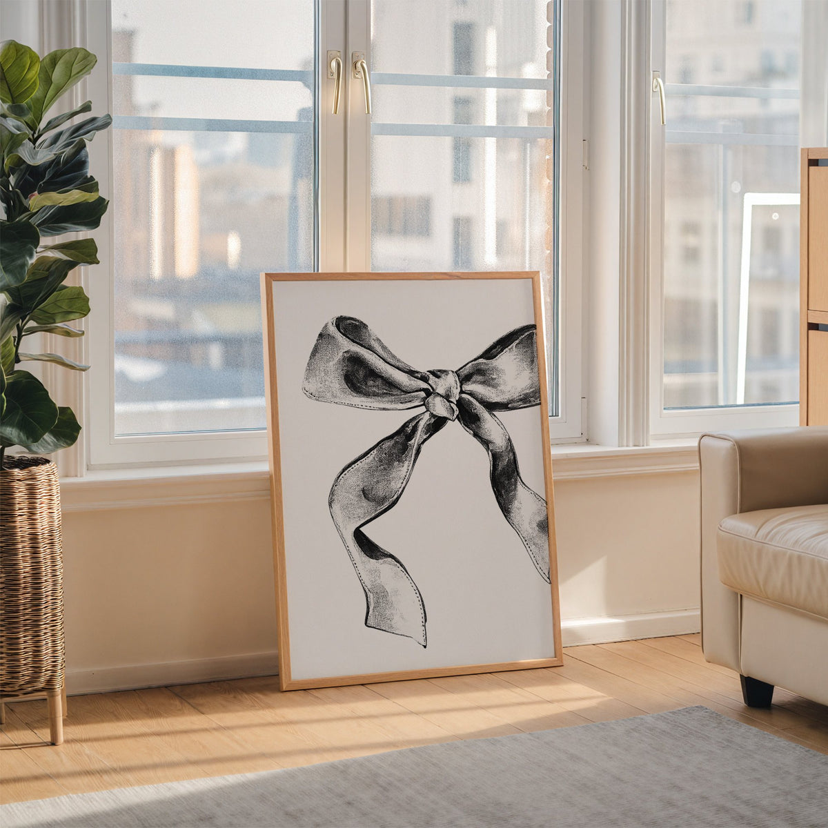 Stippled ink ribbon bow drawing print in an oak frame leaning on the floor beside a fiddle leaf fig