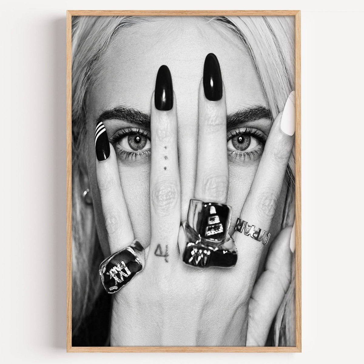 Hand with black nails and rings over eyes print in a light oak frame on a plain white wall