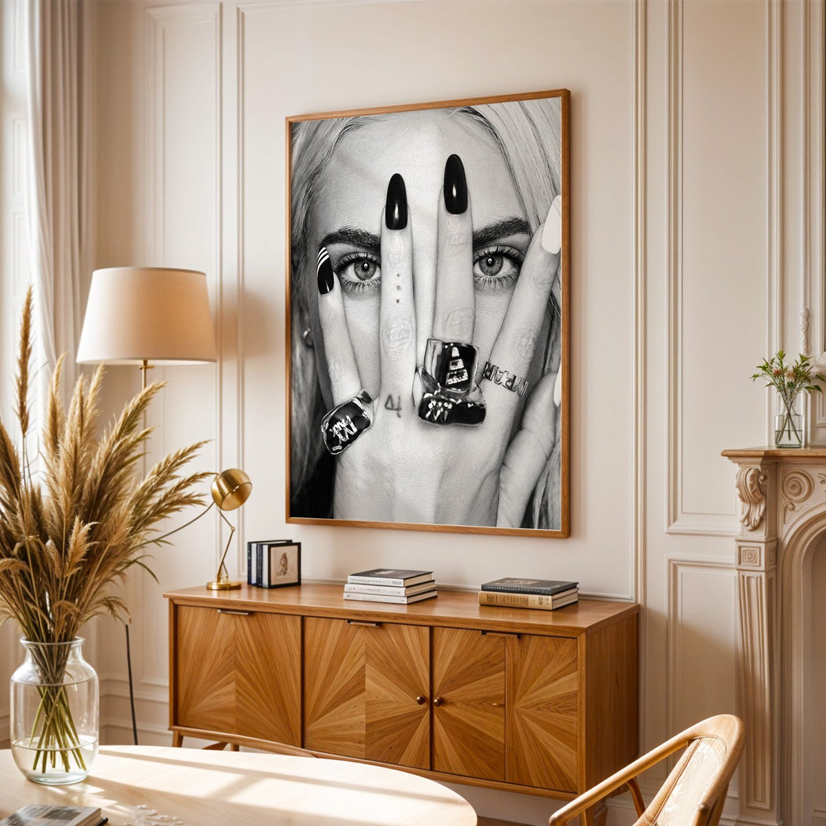 Hand with black nails and rings over eyes print in an oak frame above a sunburst wood sideboard with pampas grass