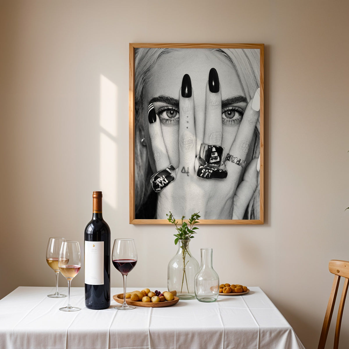 Hand with black nails and rings over eyes print in an oak frame above a table set with red wine and glasses