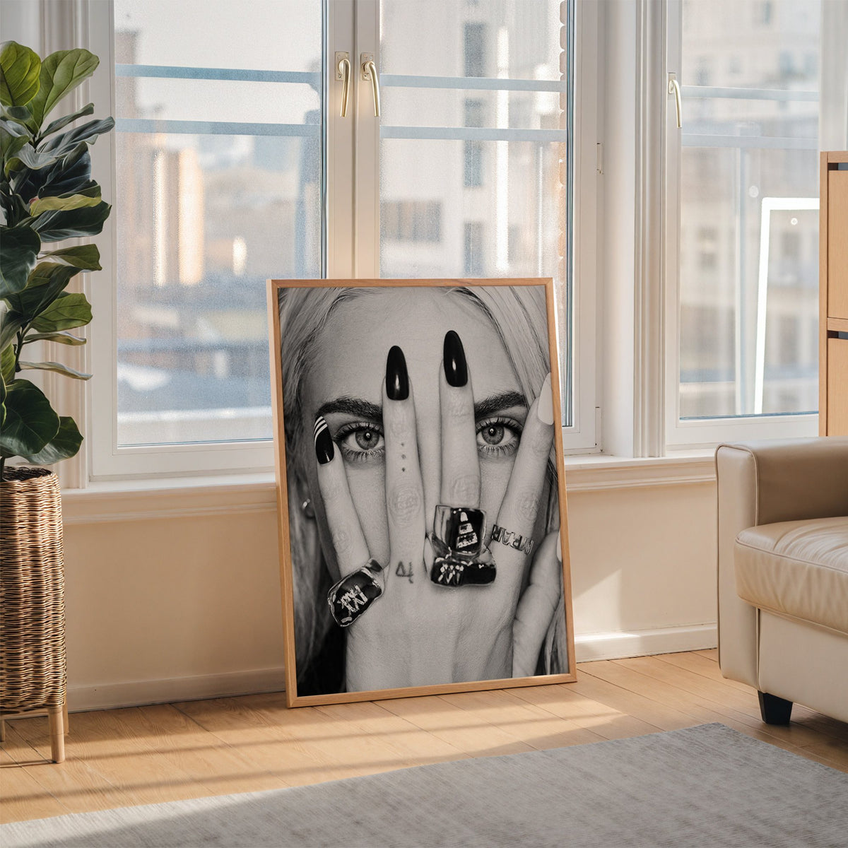 Hand with black nails and rings over eyes print in an oak frame leaning on the floor beside a fiddle leaf fig