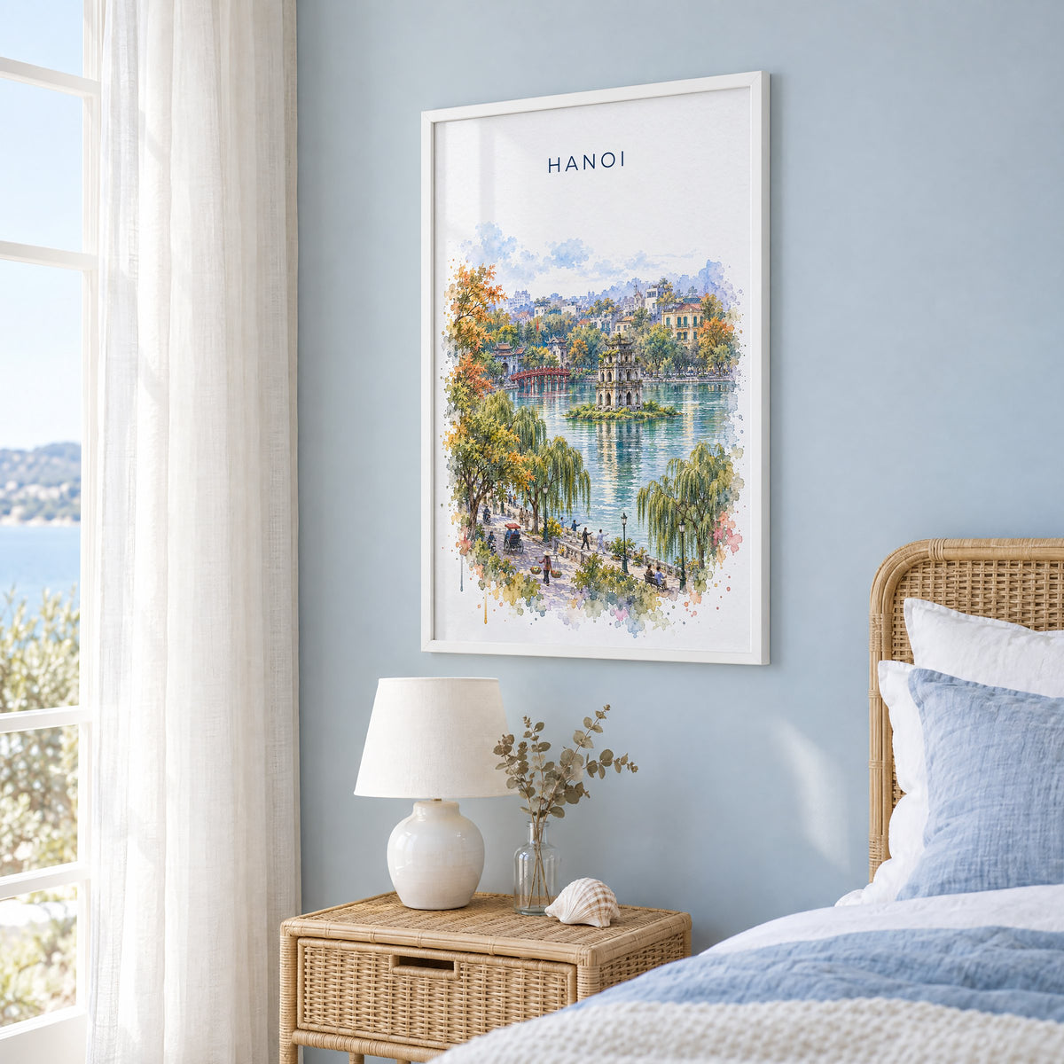Hanoi watercolor print in a white frame on a pale blue wall beside a rattan bed in a coastal bedroom