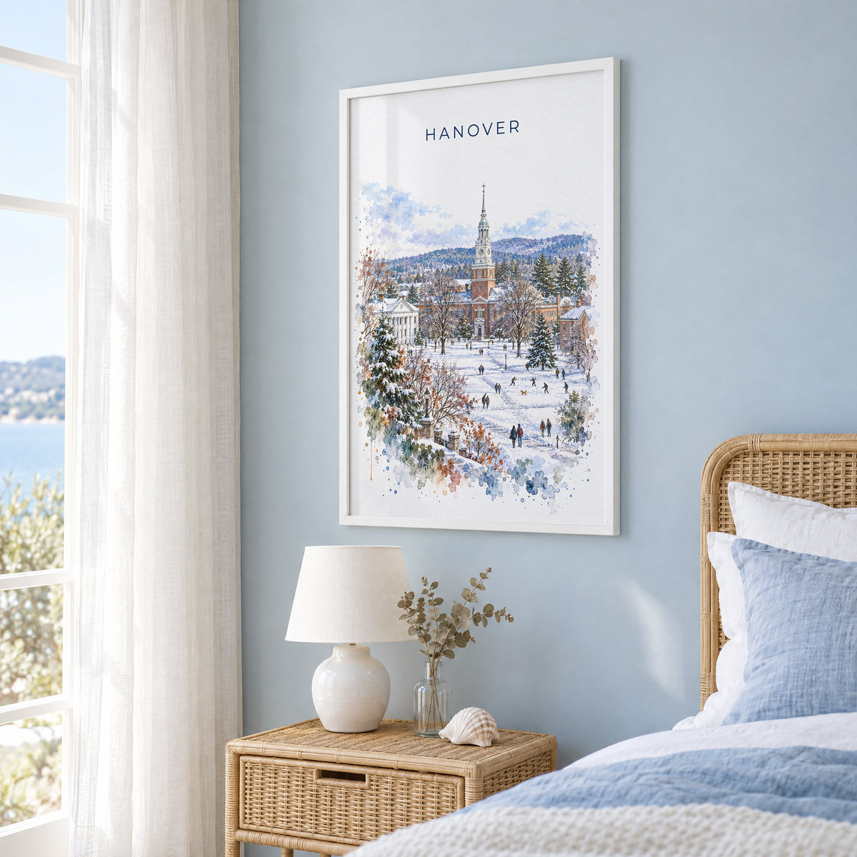 Hanover, New Hampshire watercolor print in a white frame on a pale blue wall beside a rattan bed in a coastal bedroom