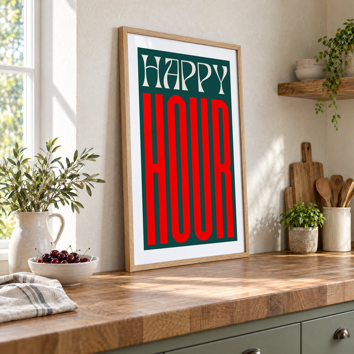 Happy Hour red and teal type print in an oak frame leaning on a butcher block island beside a bowl of cherries