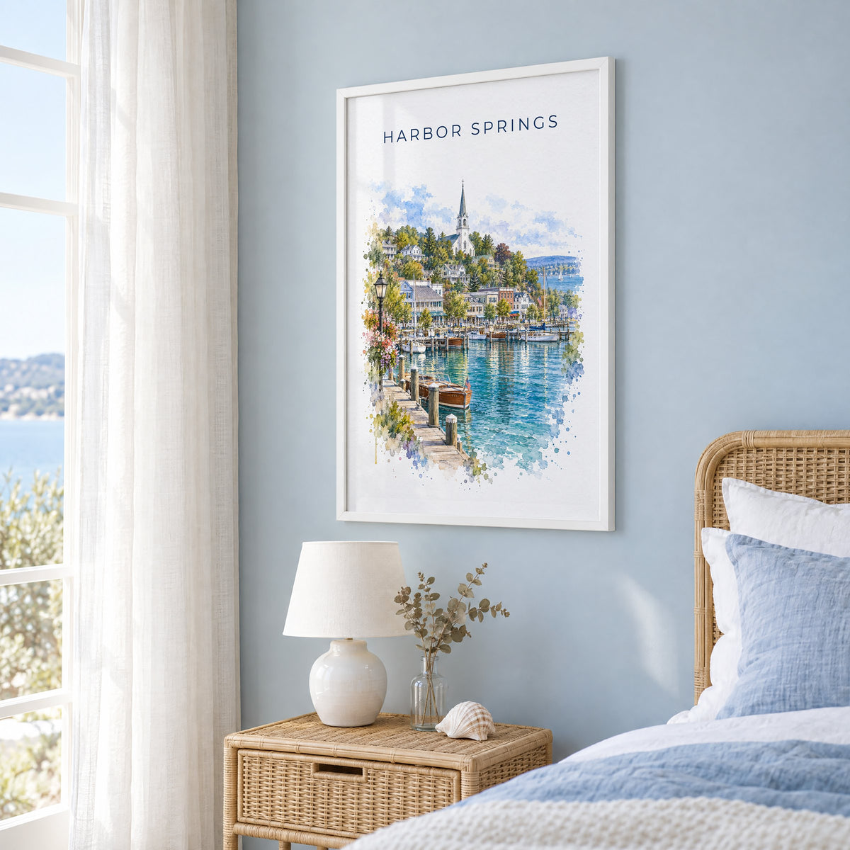 Harbor Springs, Michigan watercolor print in a white frame on a pale blue wall beside a rattan bed in a coastal bedroom