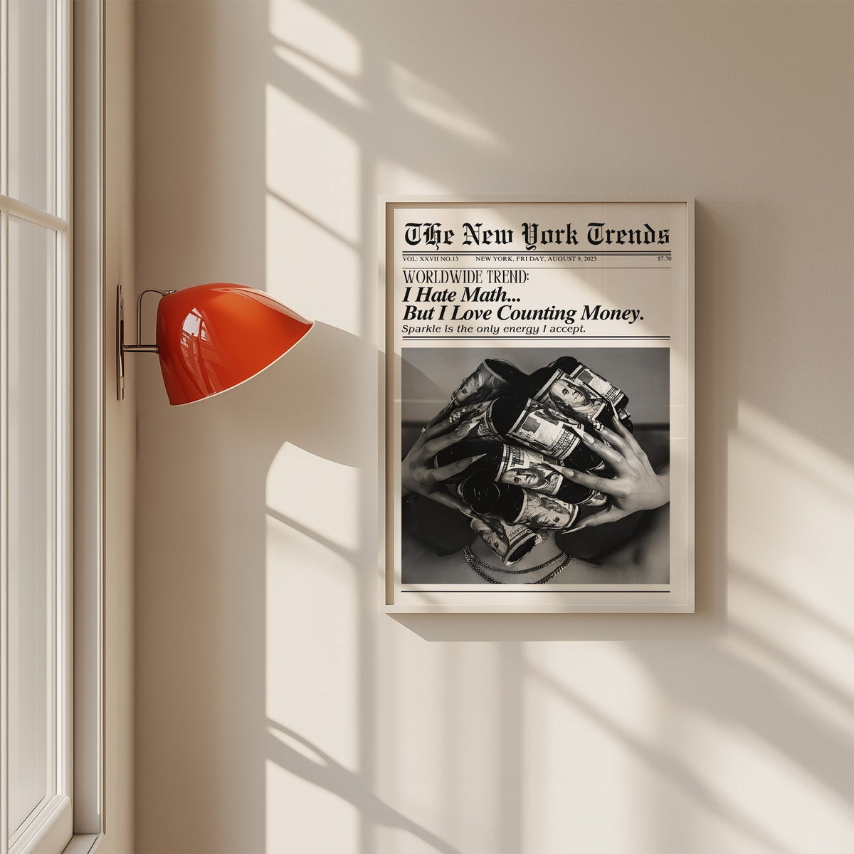 Dollar bill hair rollers mock newspaper print in a white frame on a sunlit wall beside a red sconce