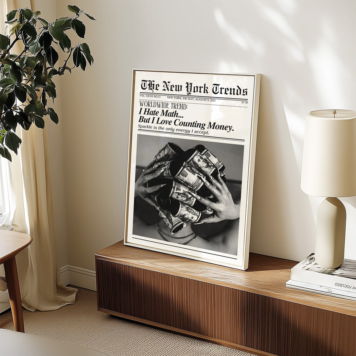 Dollar bill hair rollers mock newspaper print in a white frame on a walnut sideboard beside a ceramic lamp