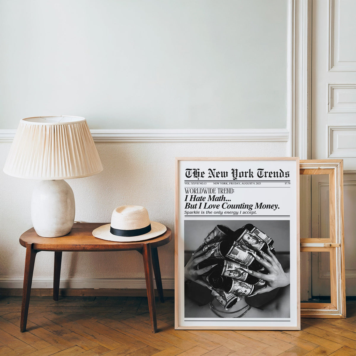 Dollar bill hair rollers mock newspaper print in a pine frame on a wood floor beside a lamp and straw hat