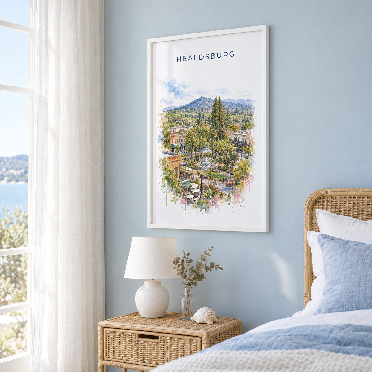Healdsburg, California watercolor print in a white frame on a pale blue wall beside a rattan bed in a coastal bedroom