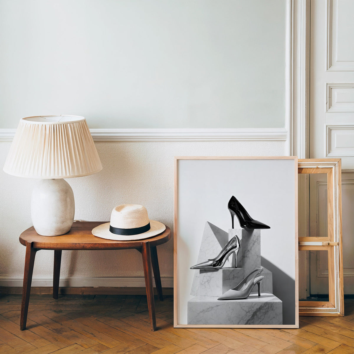 Three pairs of heels on marble pedestals print in a pine frame on a wood floor beside a lamp and straw hat