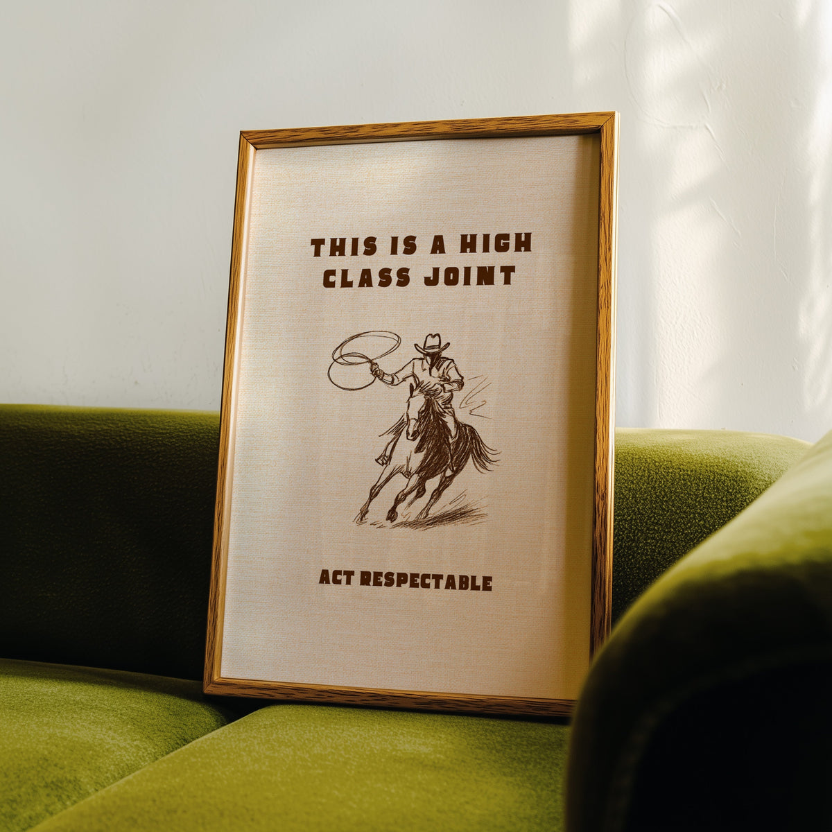 High Class Joint cowgirl cream print in an oak frame leaning on a green velvet sofa