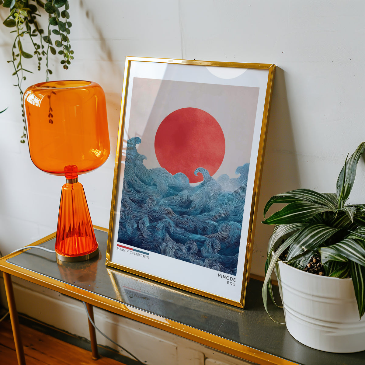 Hinode red sun over waves print shown in a gold frame on a console beside an orange glass lamp