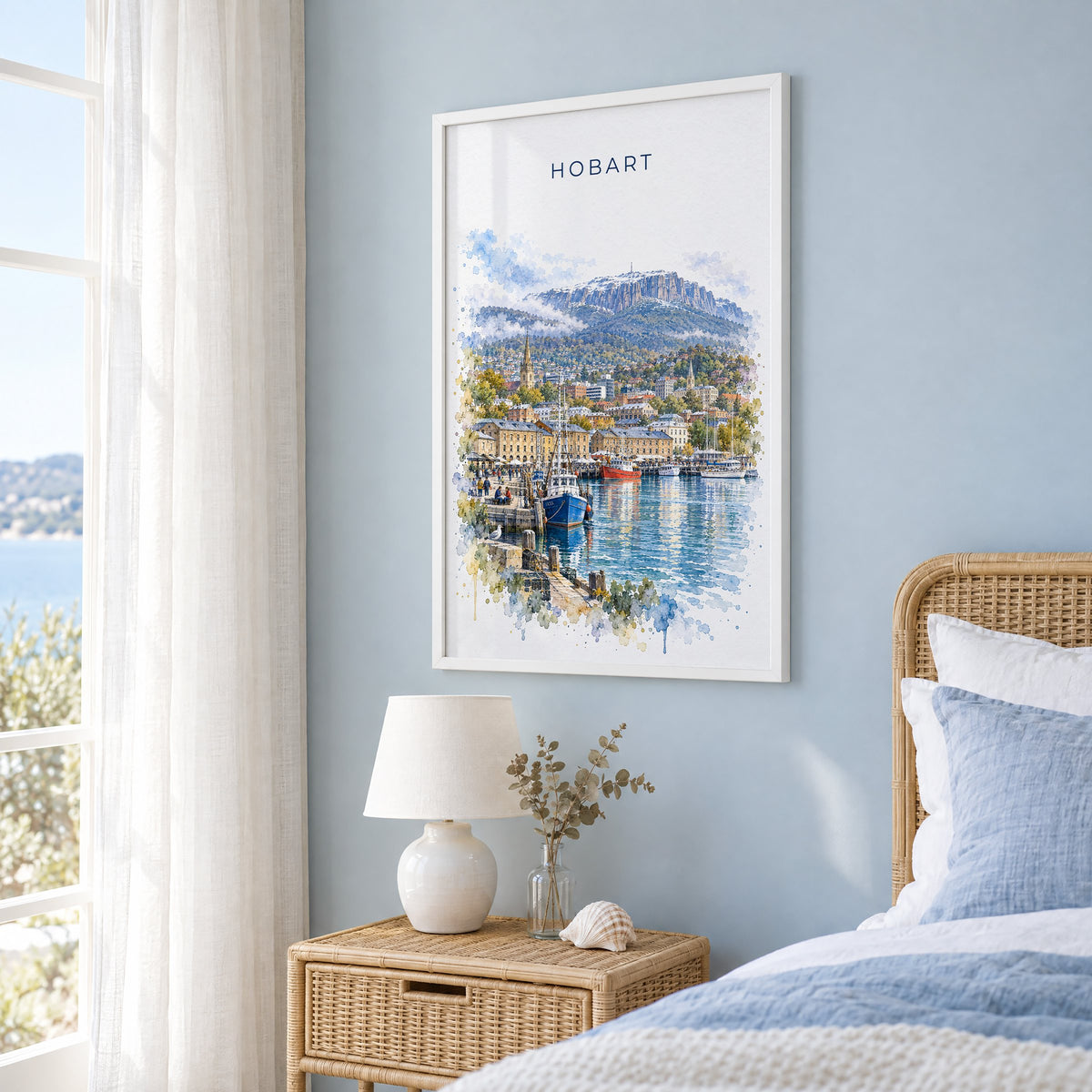 Hobart watercolor print in a white frame on a pale blue wall beside a rattan bed in a coastal bedroom