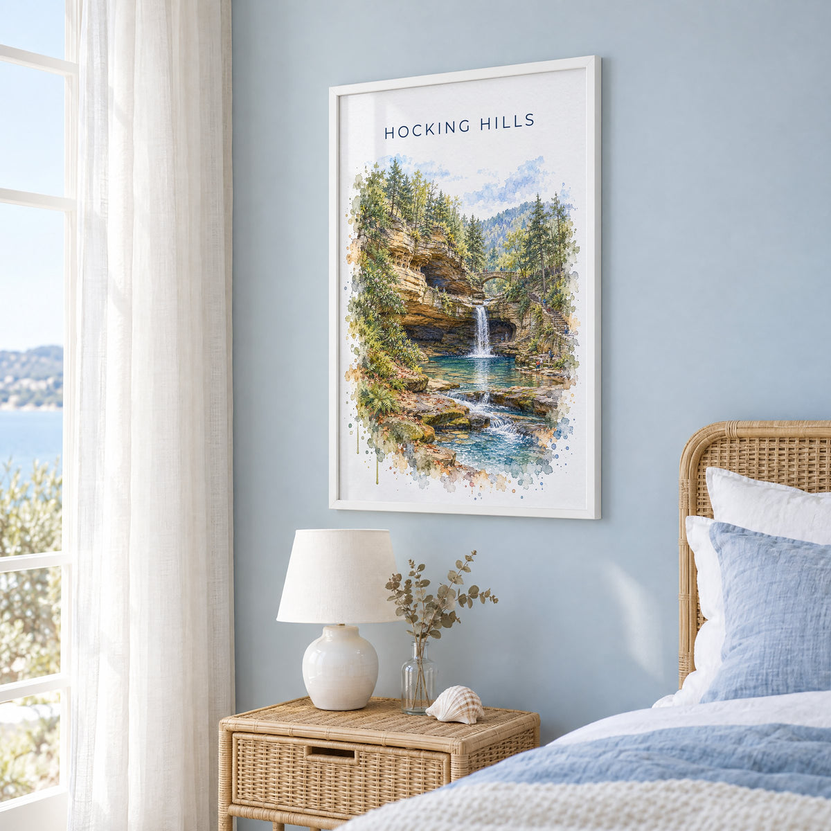 Hocking Hills watercolor print in a white frame on a pale blue wall beside a rattan bed in a coastal bedroom