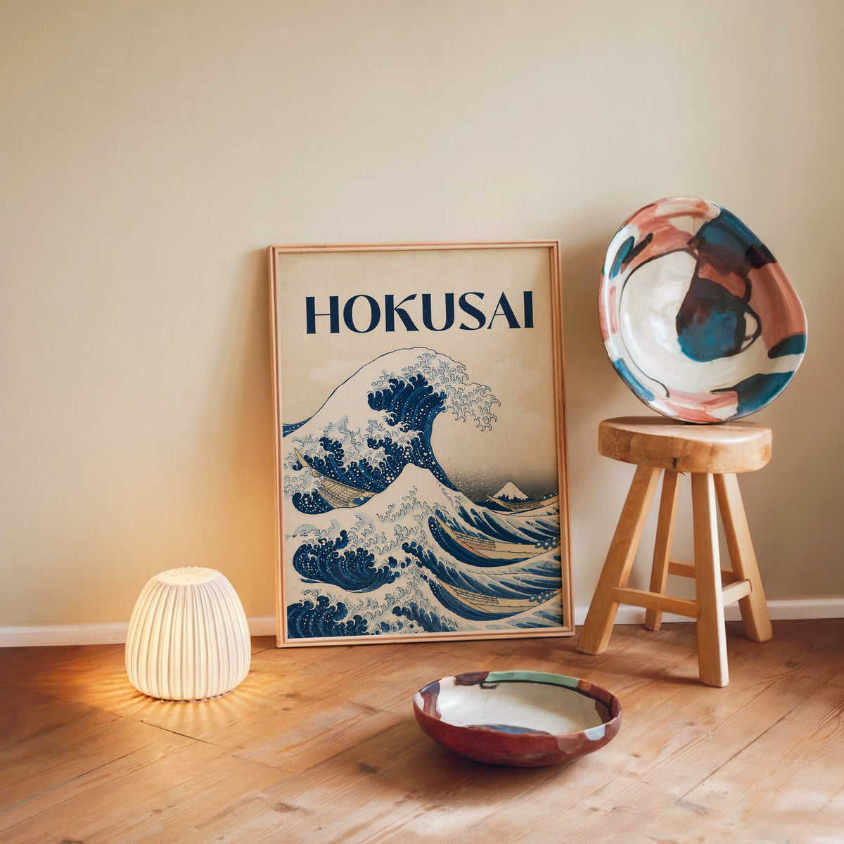 Great Wave with HOKUSAI title print in a light wood frame leaning on a wooden floor by a glowing lamp and stool