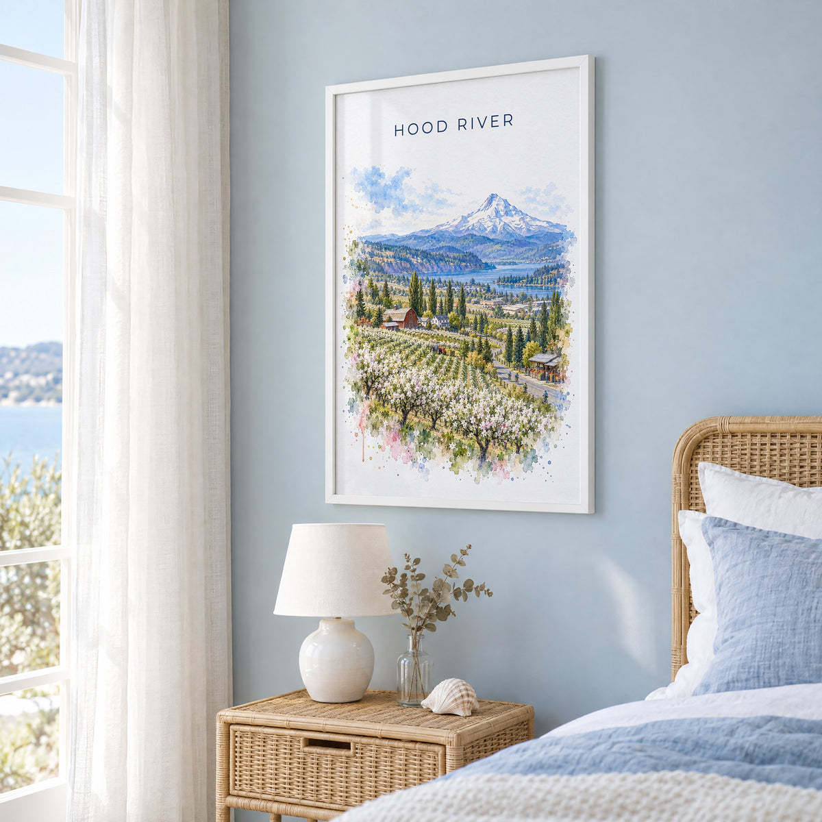 Hood River watercolor print in a white frame on a pale blue wall beside a rattan bed in a coastal bedroom