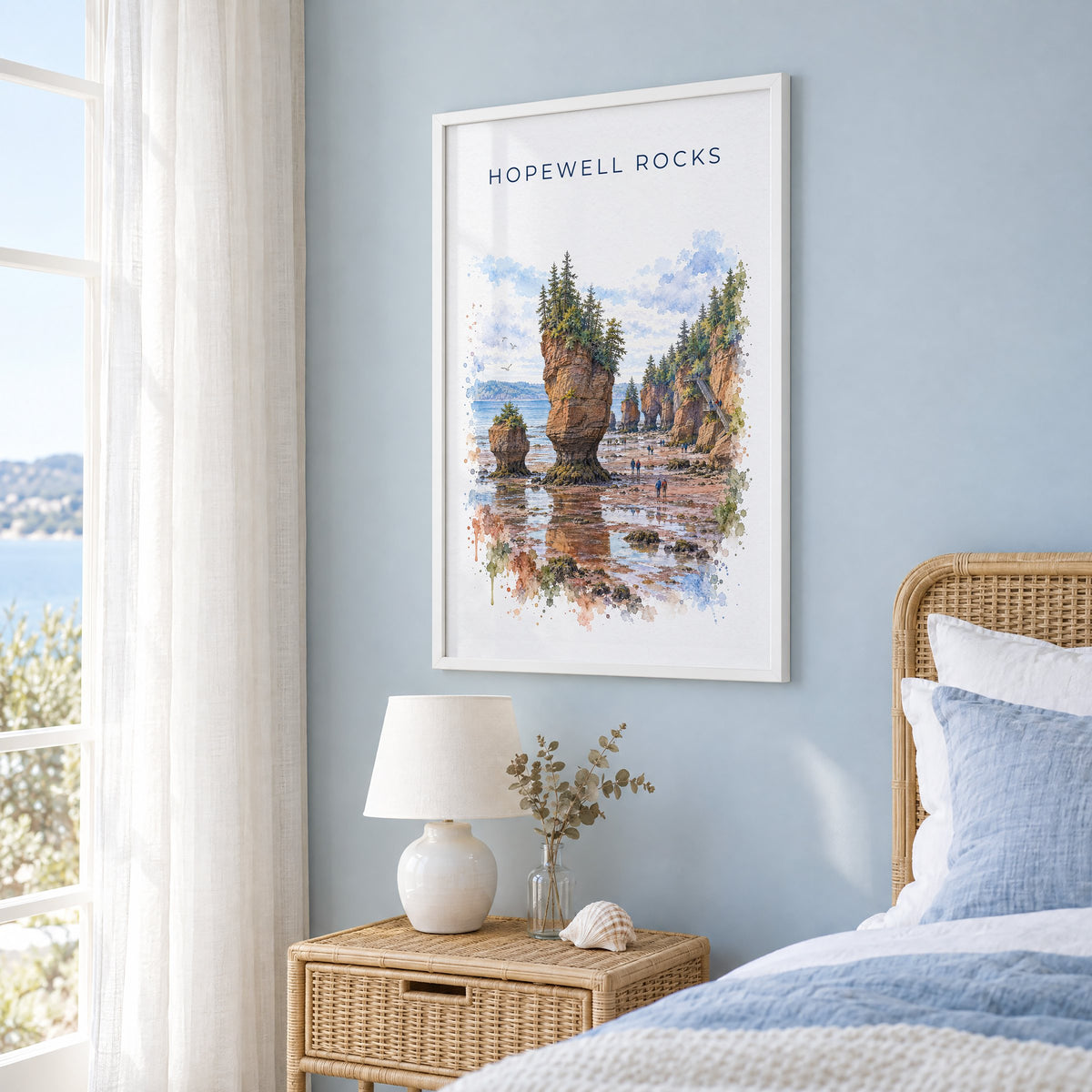 Hopewell Rocks watercolor print in a white frame on a pale blue wall beside a rattan bed in a coastal bedroom
