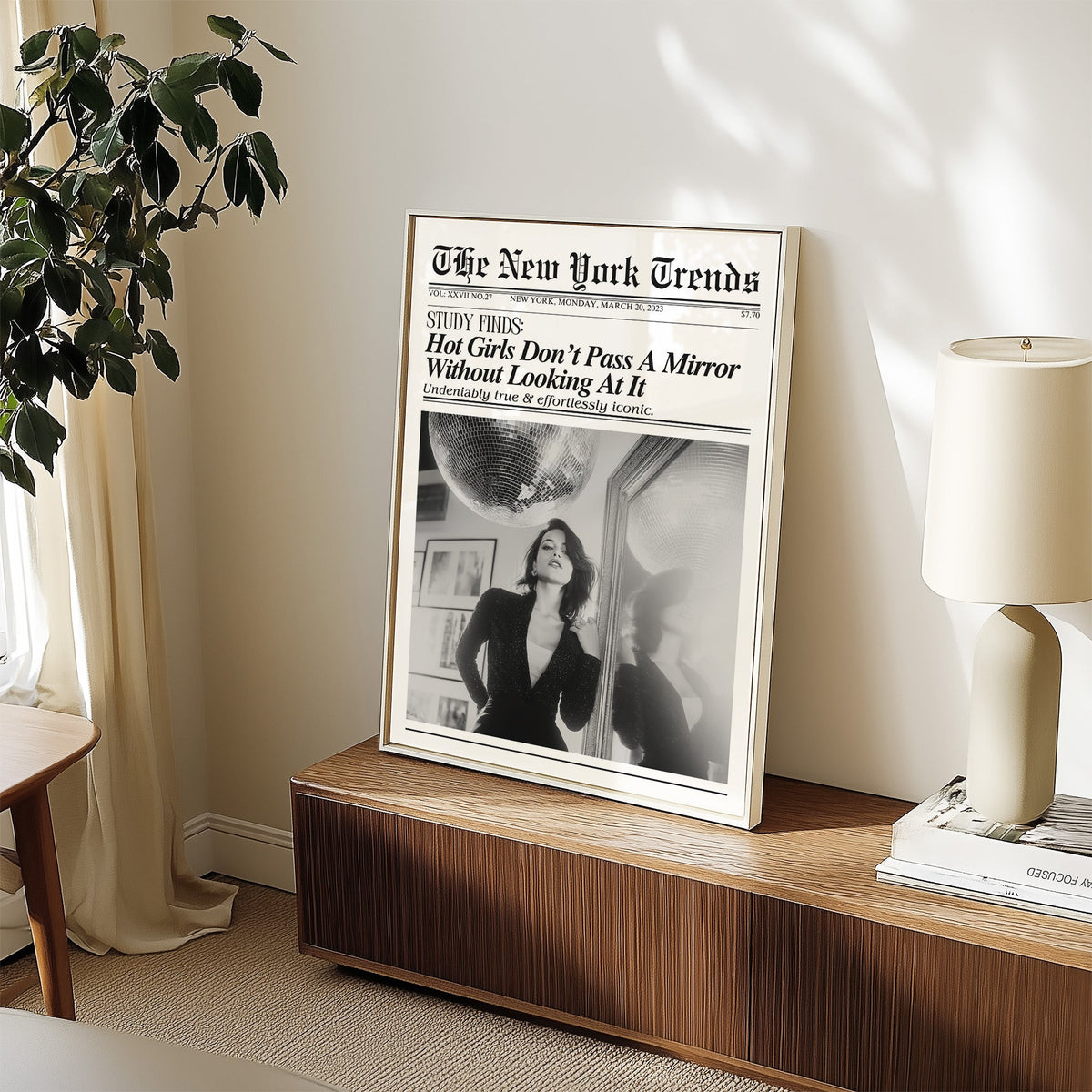 Woman by mirror with disco ball mock newspaper print in a white frame on a walnut sideboard beside a ceramic lamp