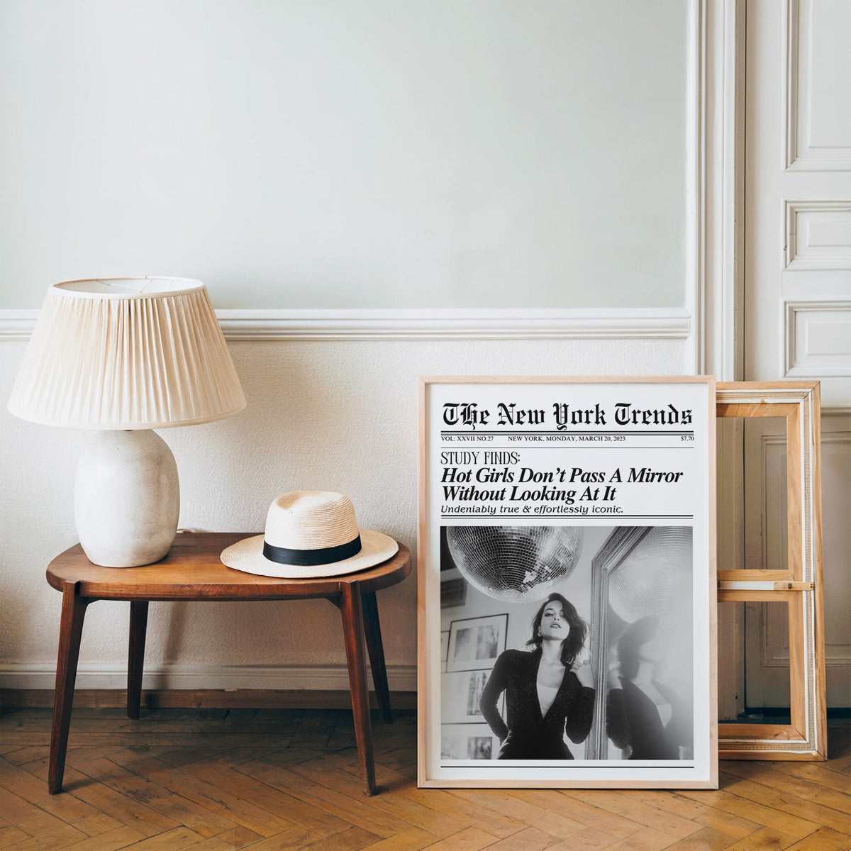 Woman by mirror with disco ball mock newspaper print in a pine frame on a wood floor beside a lamp and straw hat