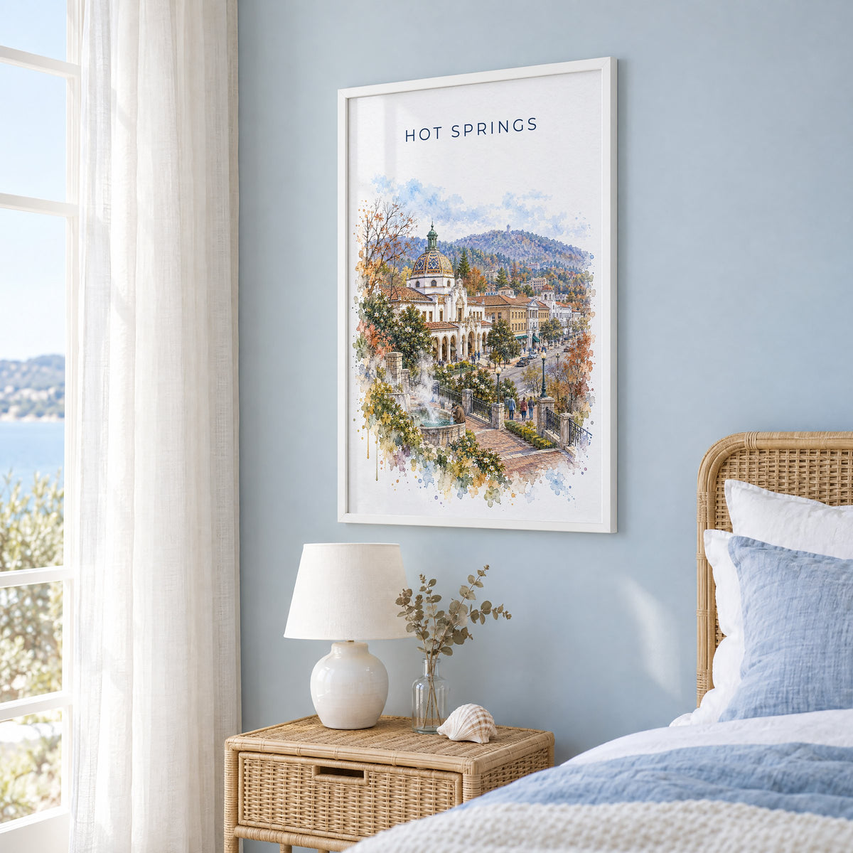 Hot Springs, Arkansas watercolor print in a white frame on a pale blue wall beside a rattan bed in a coastal bedroom