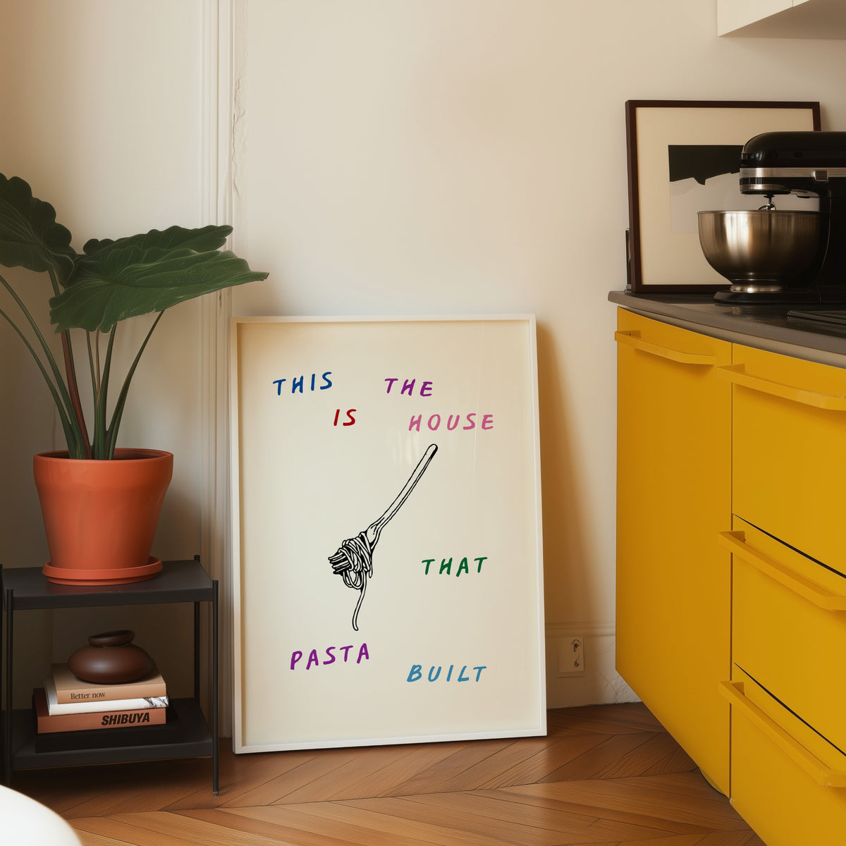 House That Pasta Built print in a white frame on parquet beside a yellow kitchen