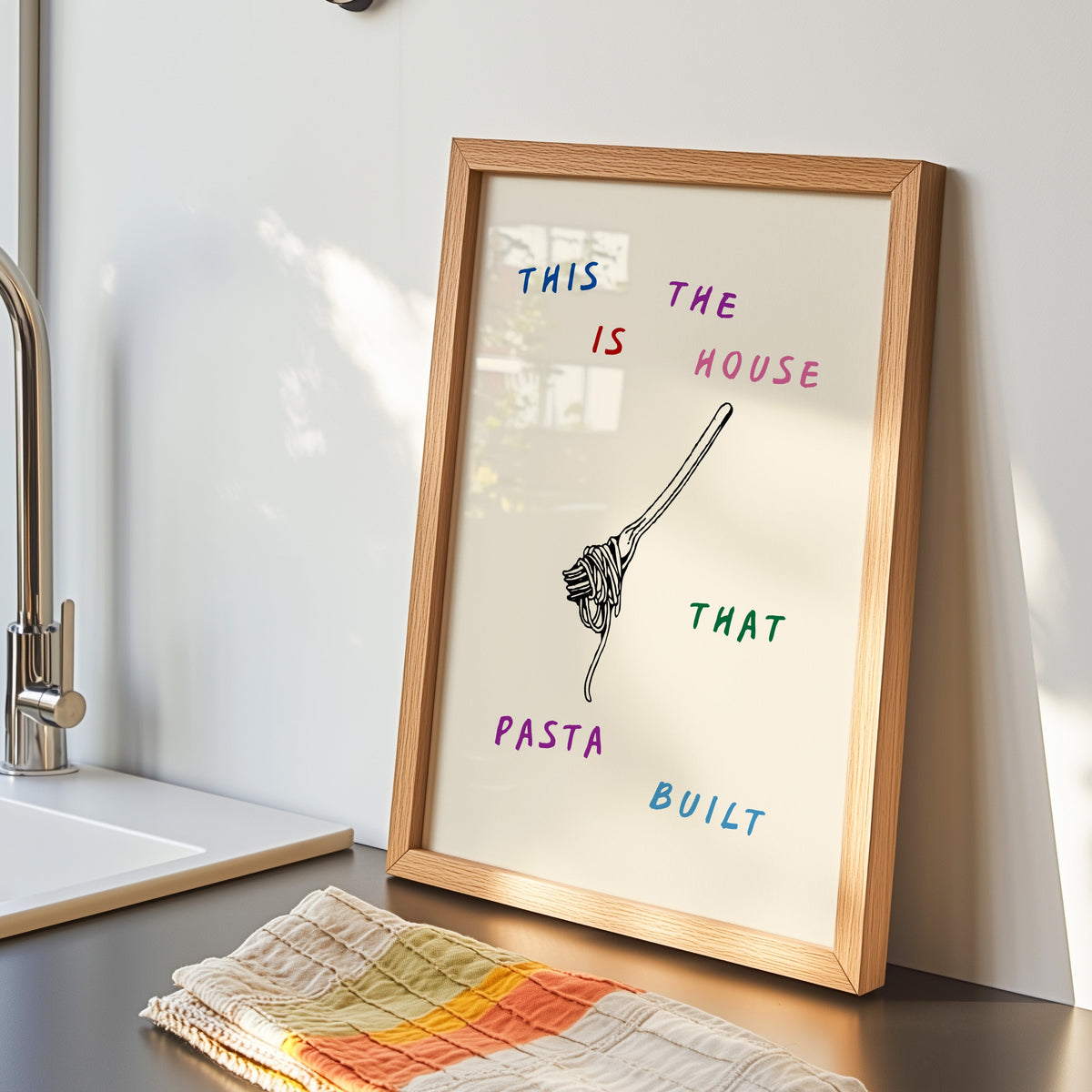 House That Pasta Built print in an oak frame by the kitchen sink with a tea towel