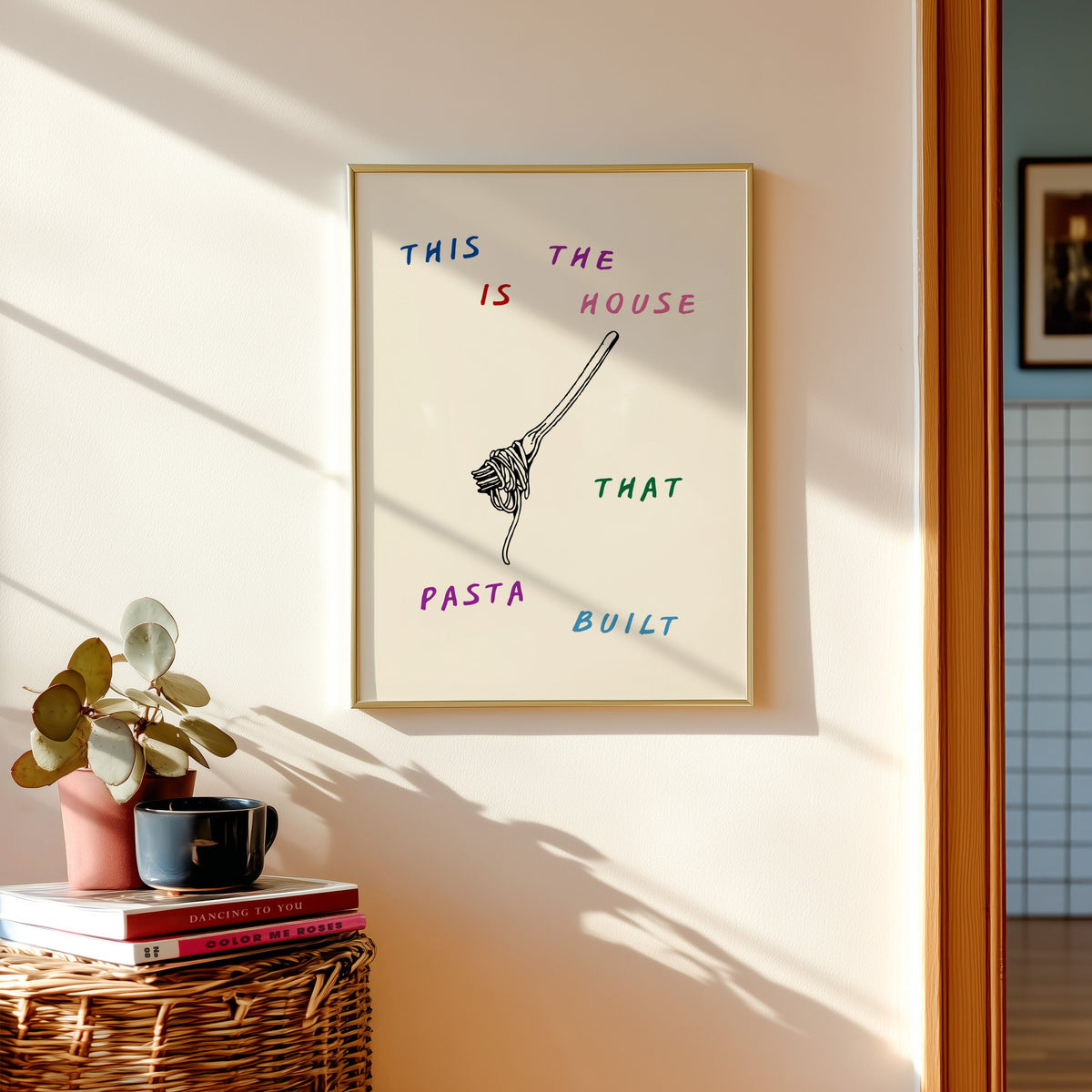 House That Pasta Built print in a gold frame above a basket of books and a pilea plant
