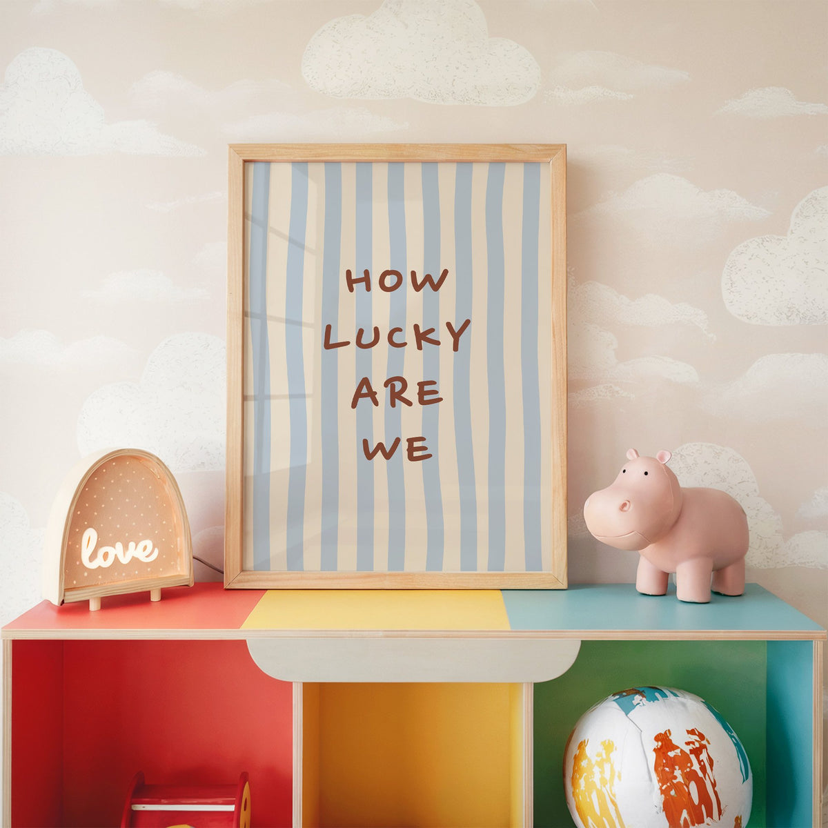How lucky are we nursery print in an oak frame on a rainbow toy shelf against cloud wallpaper