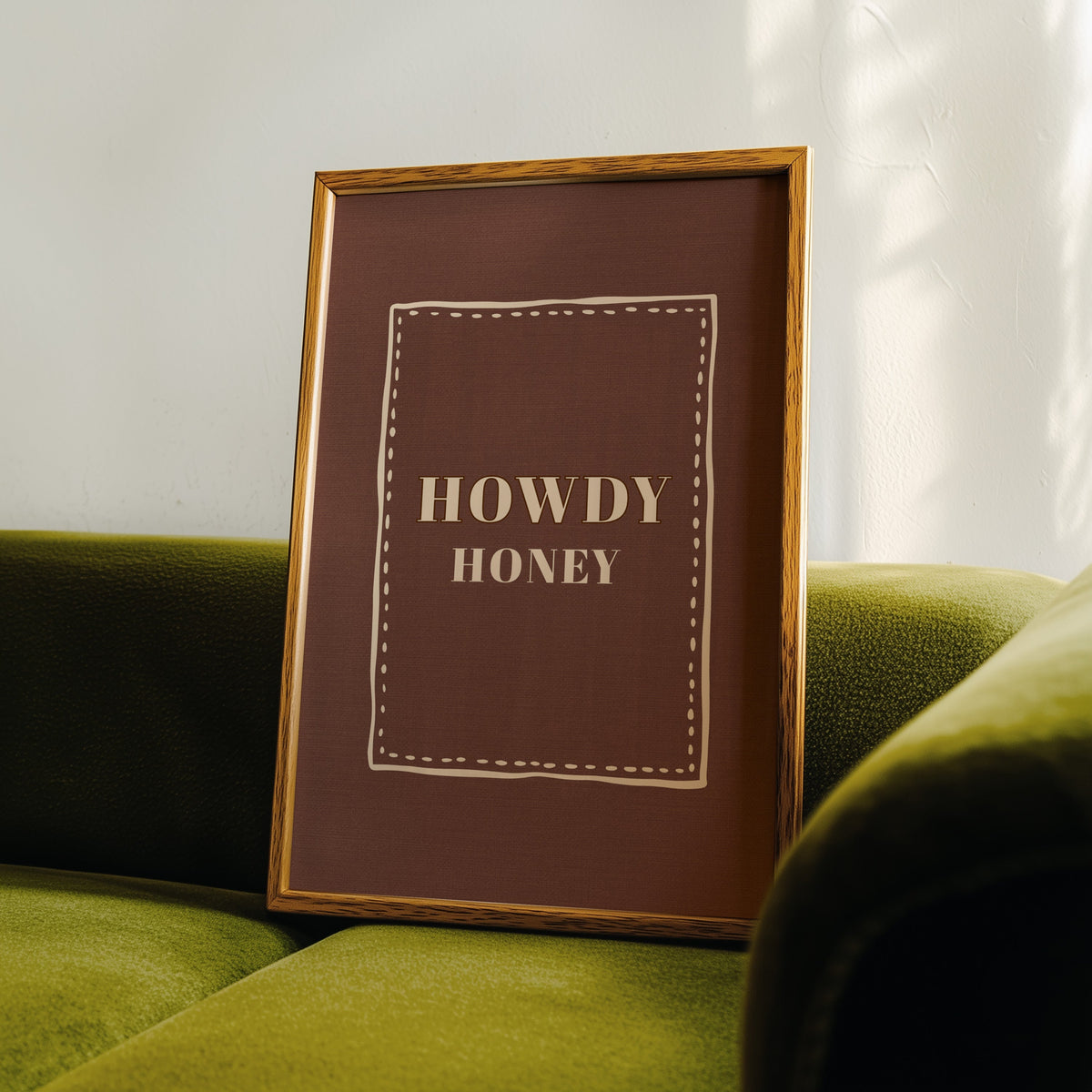 Howdy Honey brown print in an oak frame leaning on a green velvet sofa