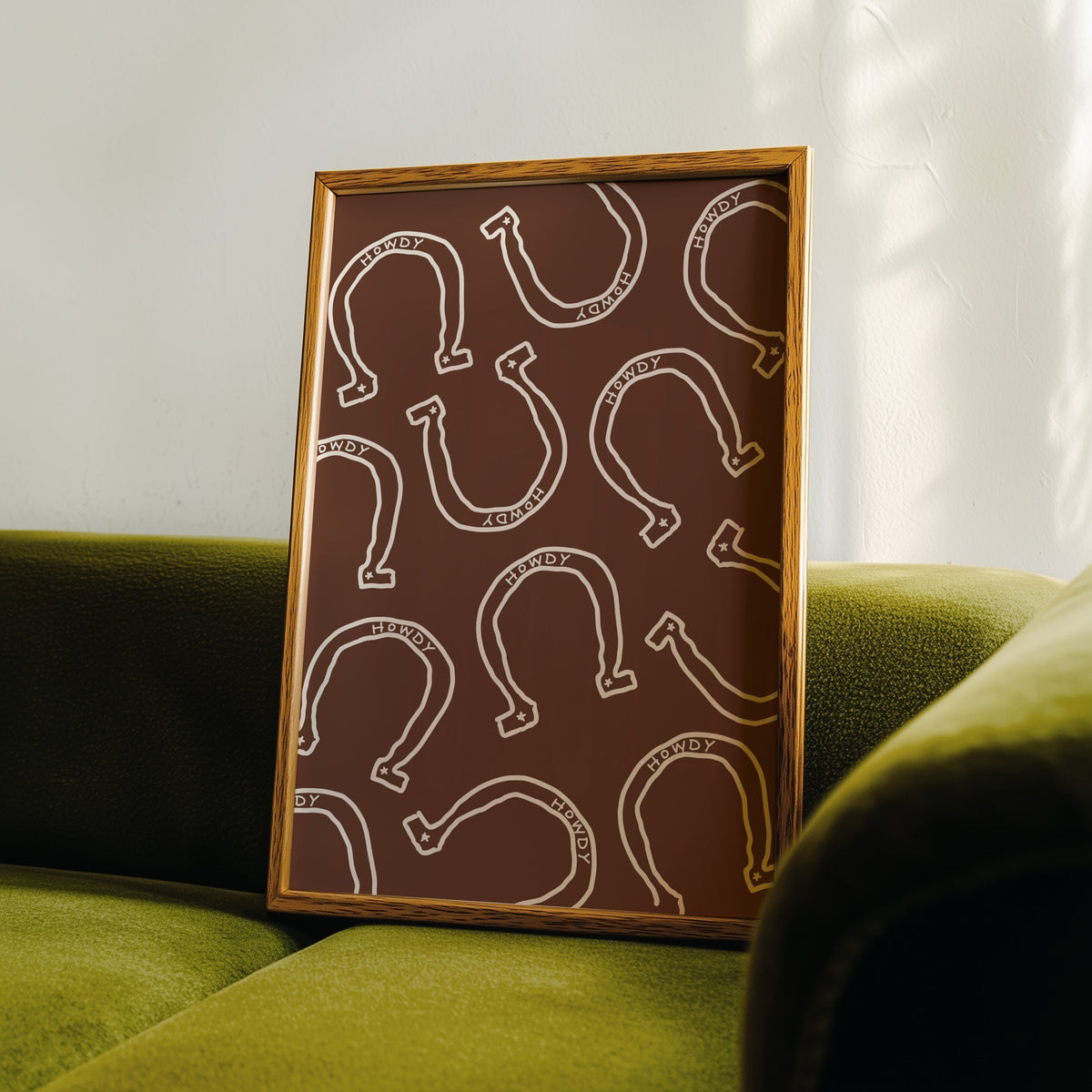 Howdy horseshoe brown pattern print in an oak frame leaning on a green velvet sofa