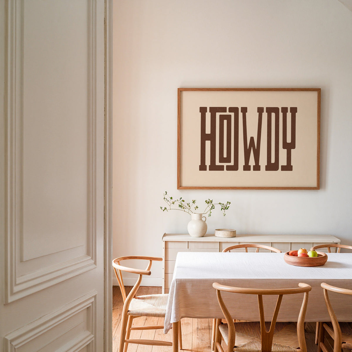 Howdy slab serif cream print in an oak frame above a dining table with wishbone chairs