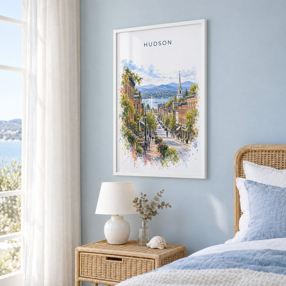 Hudson, New York watercolor print in a white frame on a pale blue wall beside a rattan bed in a coastal bedroom