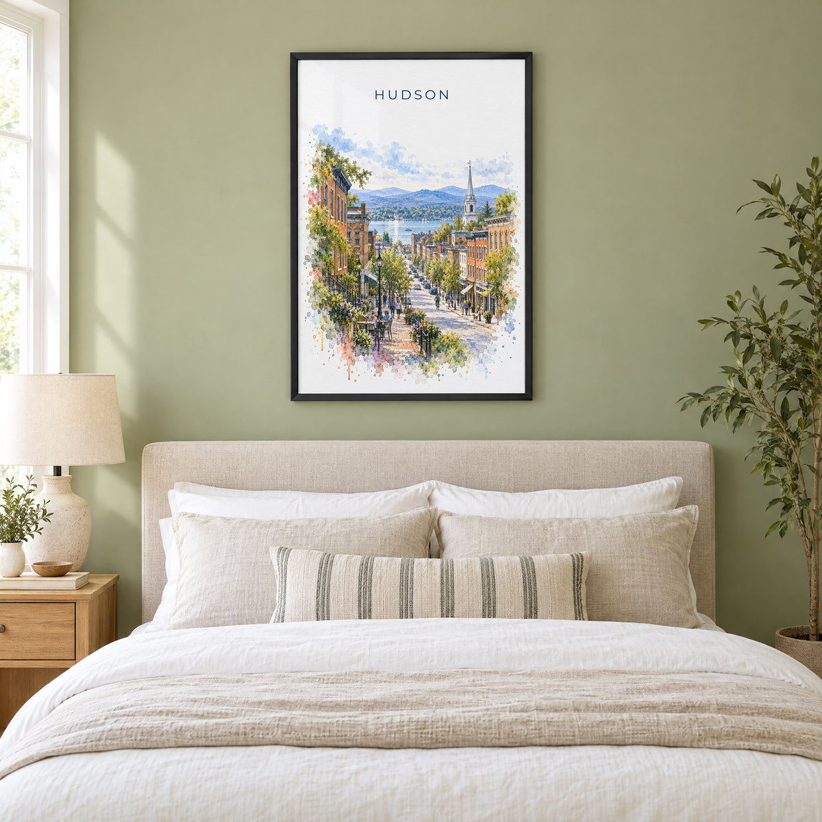 Hudson, New York watercolor print in a black frame above an upholstered headboard in a sage green bedroom