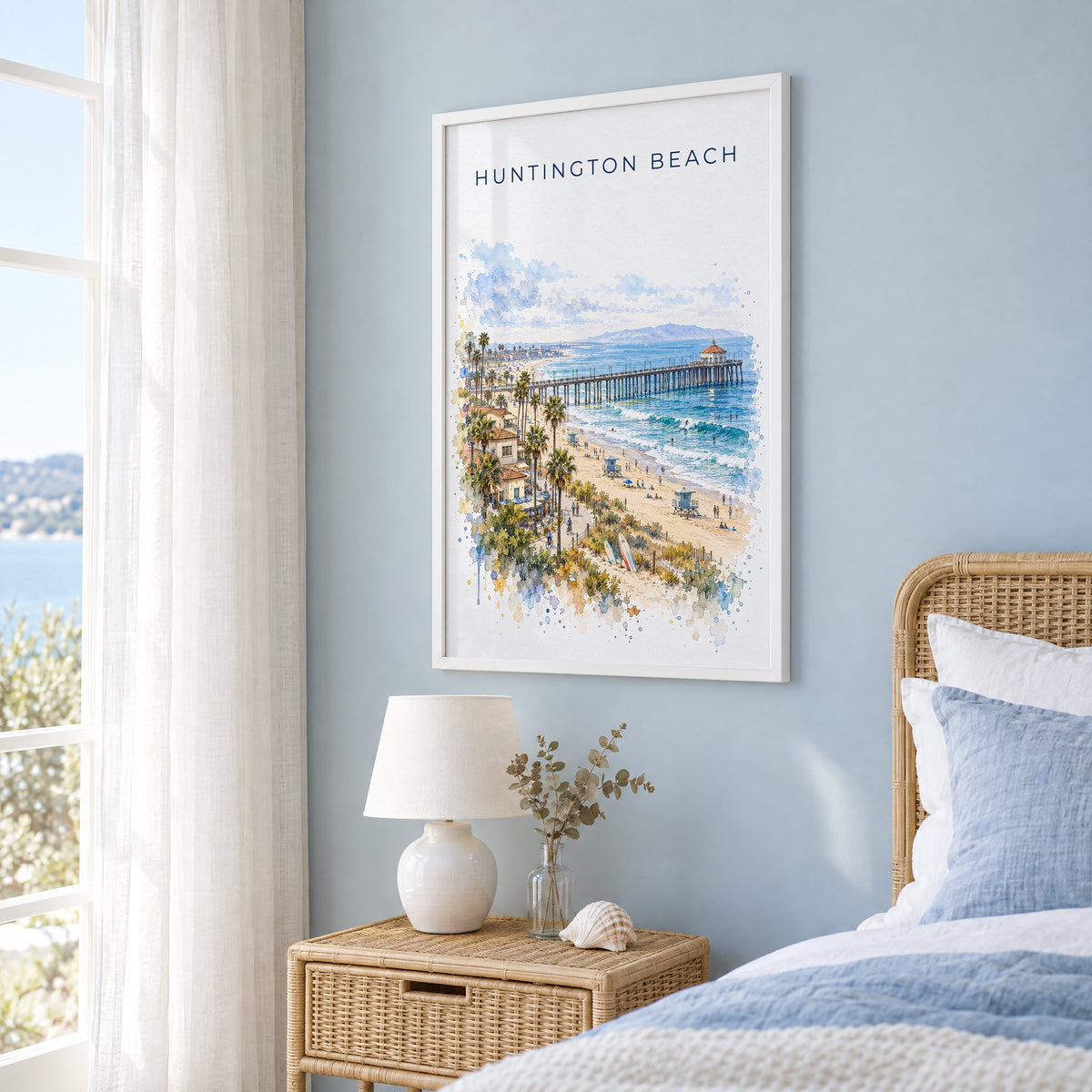 Huntington Beach watercolor print in a white frame on a pale blue wall beside a rattan bed in a coastal bedroom