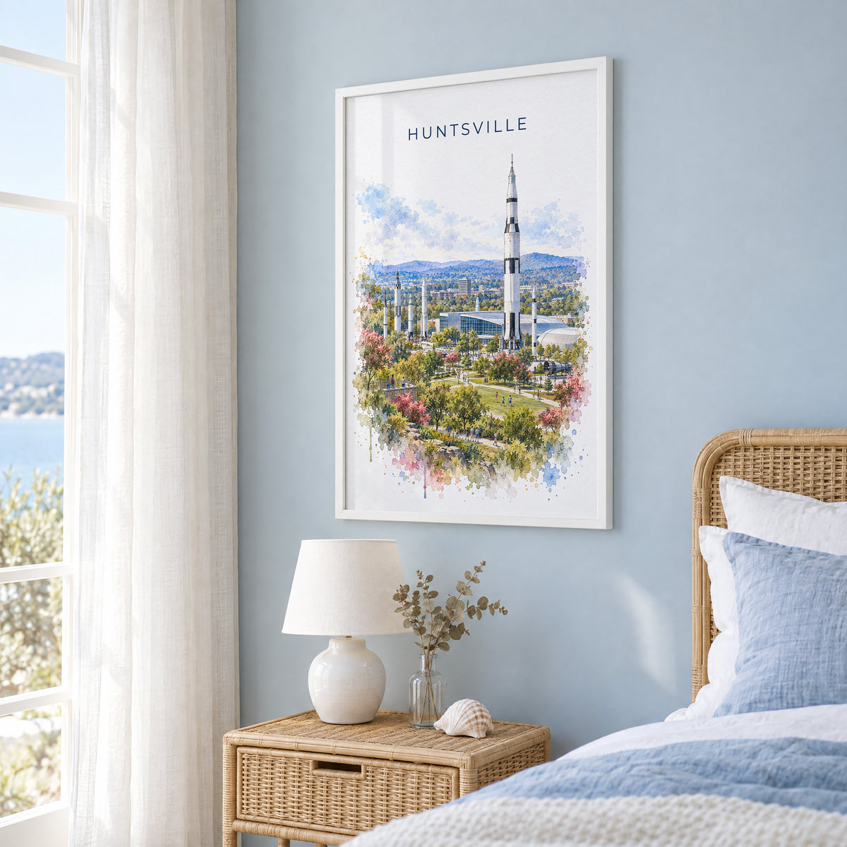 Huntsville watercolor print in a white frame on a pale blue wall beside a rattan bed in a coastal bedroom