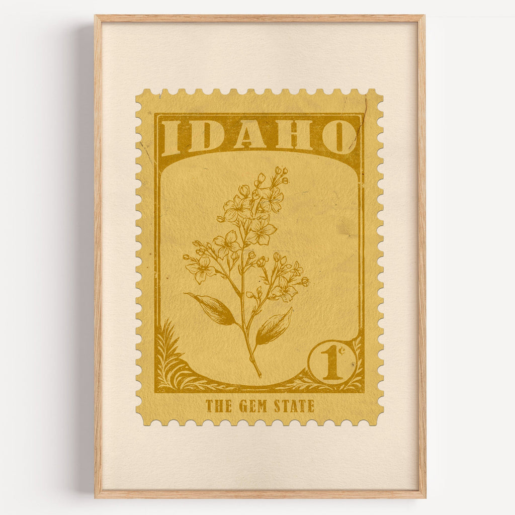 Idaho state flower stamp print in mustard gold on wheat, light oak frame on a plain white wall