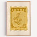 Idaho state flower stamp print in mustard gold on wheat, light oak frame on a plain white wall