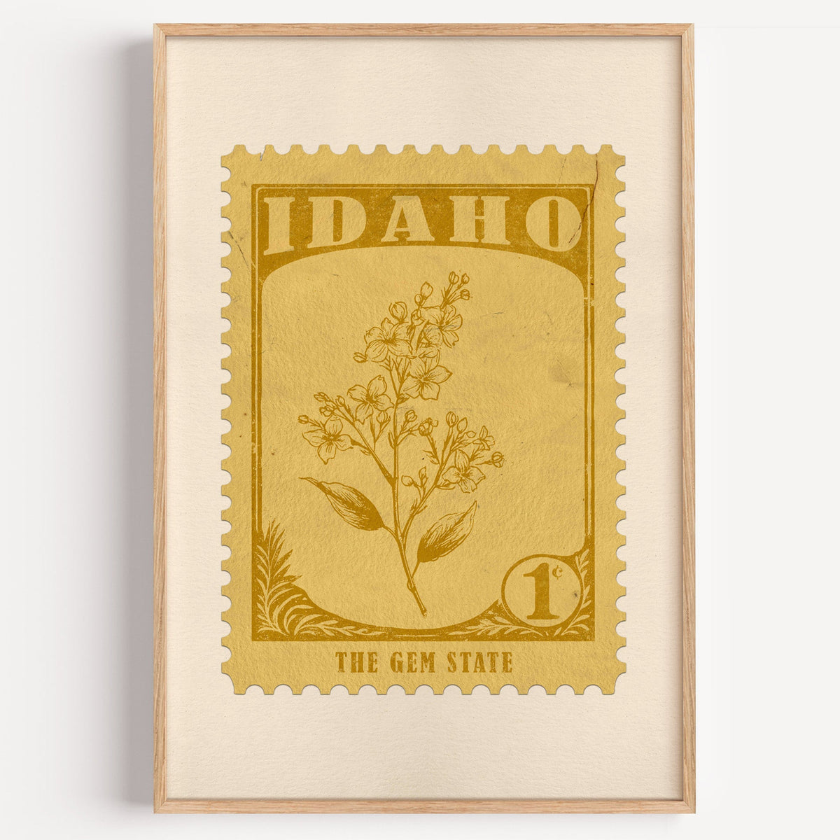 Idaho state flower stamp print in mustard gold on wheat, light oak frame on a plain white wall