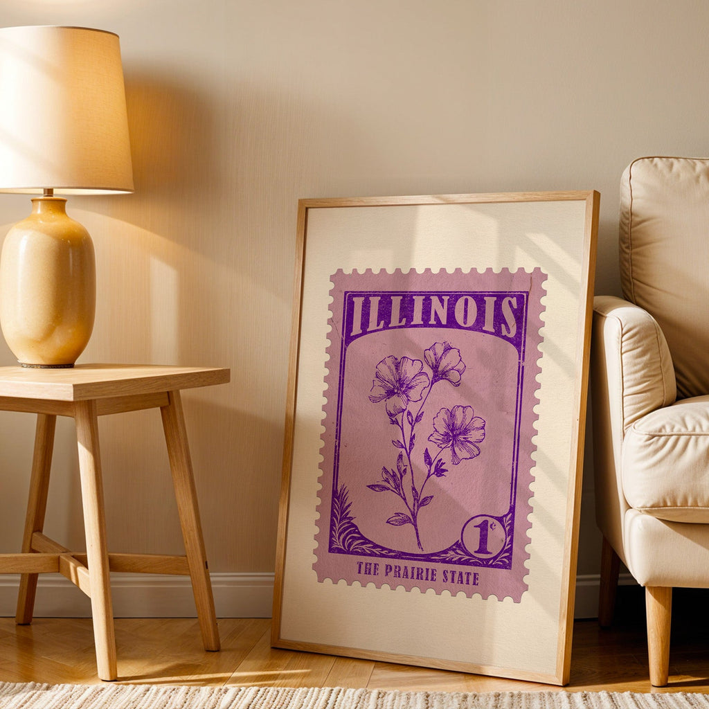 Illinois stamp print in a wood frame resting on the floor beside a beige armchair and a yellow lamp
