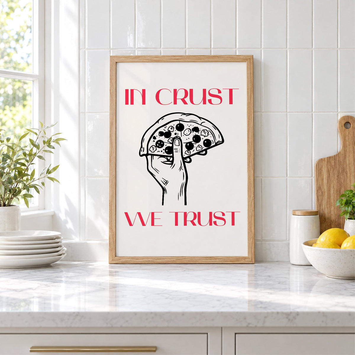 In Crust We Trust pizza print in an oak frame leaning on a marble counter by white tiles and lemons