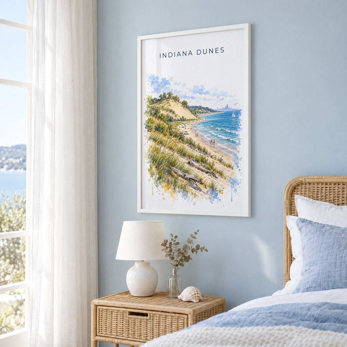 Indiana Dunes National Park watercolor print in a white frame on a pale blue wall beside a rattan bed in a coastal bedroom