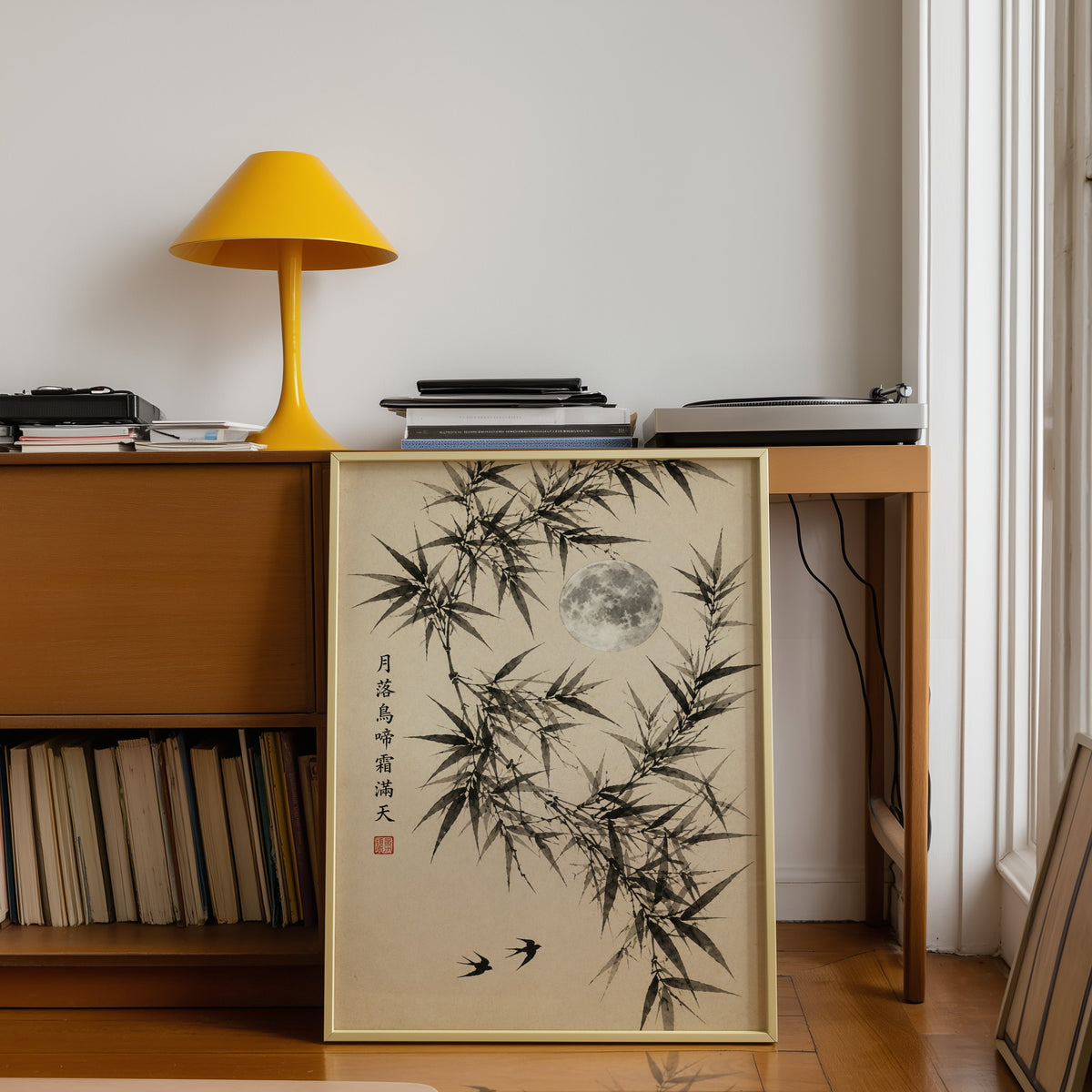 ink bamboo, full moon and swallows print shown in a gold frame leaning under a teak desk with a yellow lamp