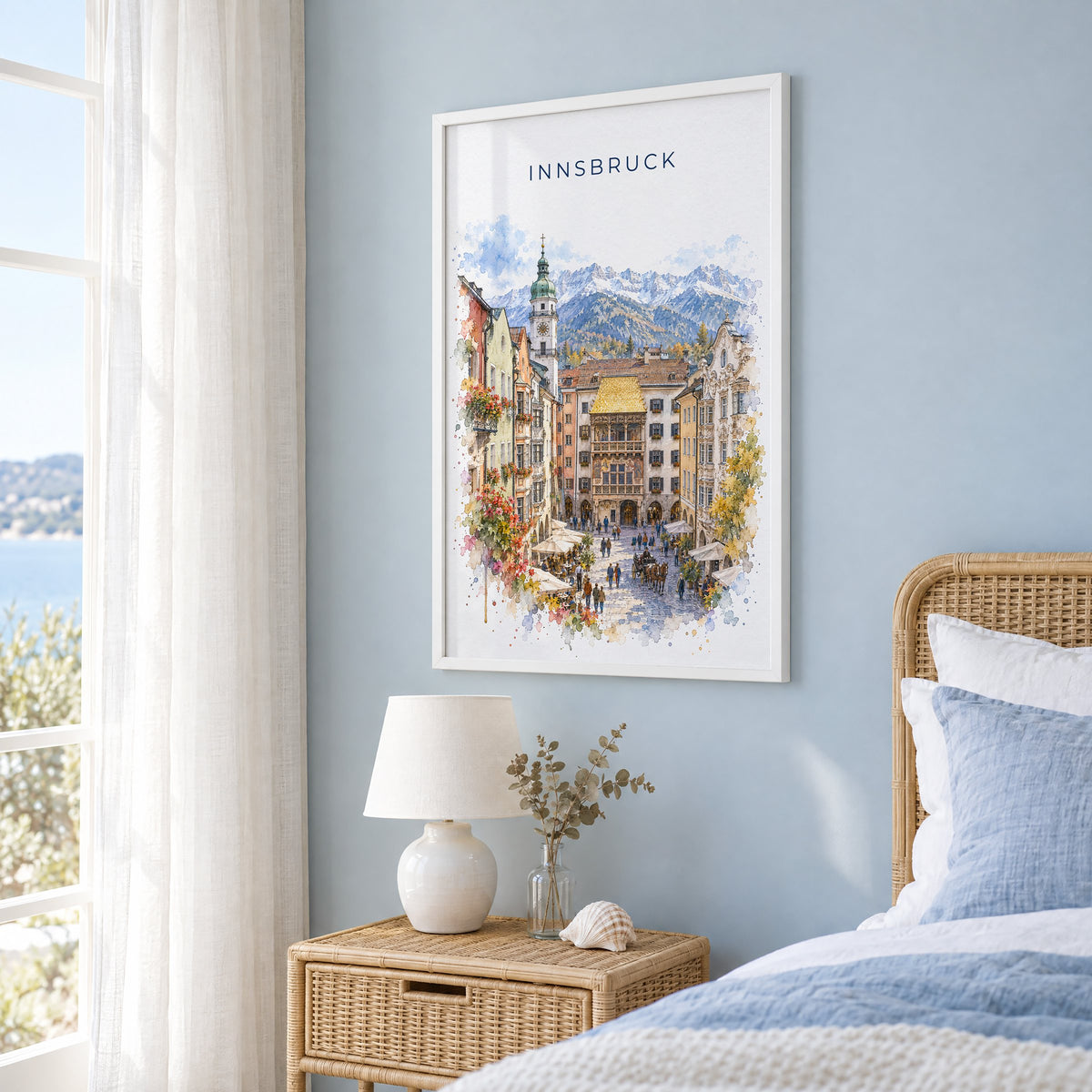Innsbruck watercolor print in a white frame on a pale blue wall beside a rattan bed in a coastal bedroom