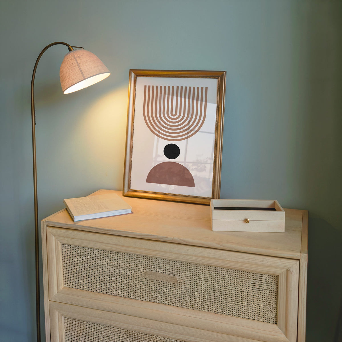 Inverted tan arch and mauve dome mid century print shown in a gold frame on a cane dresser lit by a floor lamp